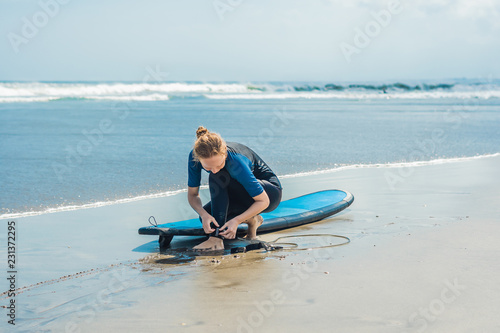 Summer time and active rest concept. Young surfer woman beginner fastens leash across leg, going to surf on big barral waves on ocean, dresseed in boardies, poses aganist sky on hot sunny day photo