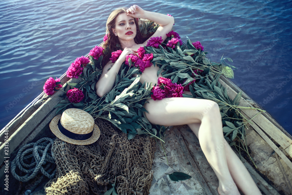 Beautiful woman with perfect body shape lying in the wooden boat covered  with peonies. Fishing net and straw hat on the bottom next to her. Deep  blue water around. Stock Photo