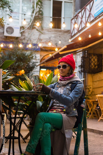 Young hipster beautiful woman (girl) sitting in cozy cafe terrace at table making smartphone selfie. Red hat and big shades. Lifestyle concept. Traveling tourist hipster in Georgia, Asia.