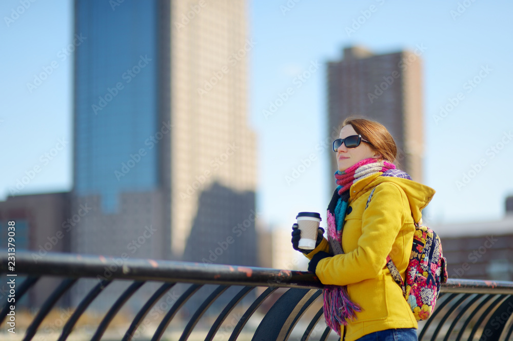 Fototapeta premium Happy young woman tourist sightseeing in New York City at sunny spring day. Female traveler drinking coffee in downtown Manhattan.