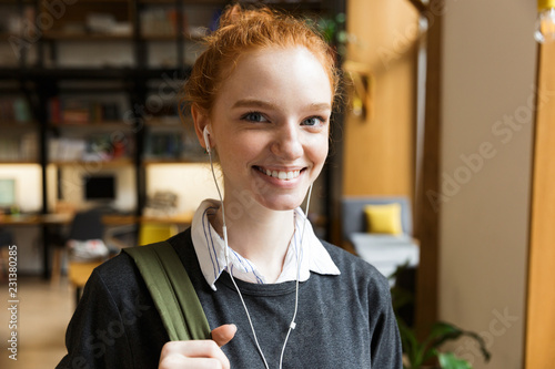 Redhead lady student posing indoors in library istening music with earphones. photo