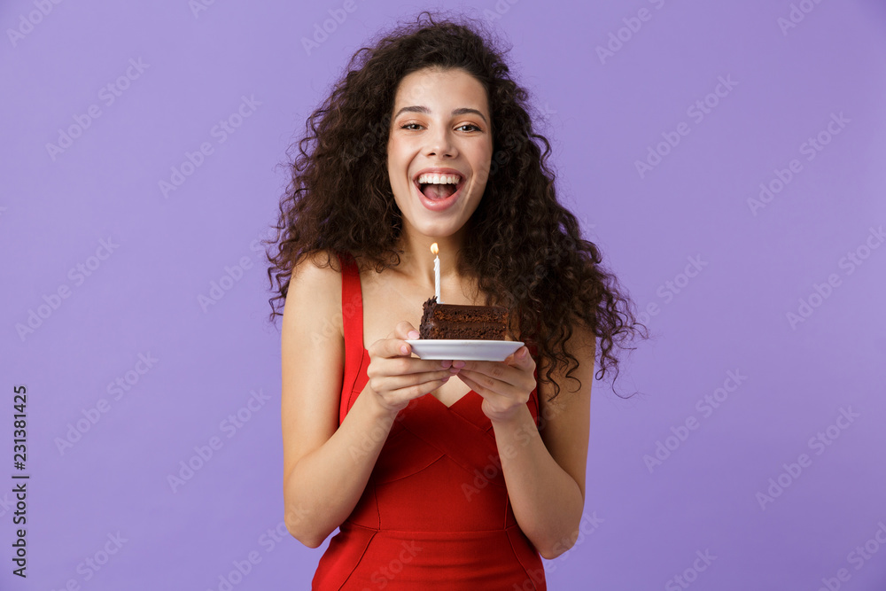 Image of happy woman 20s wearing red dress holding piece of birthday cake with candle, isolated over violet background Image of happy woman 20s wearing red dress holding piece of birthday cake with candle, isolated over violet background