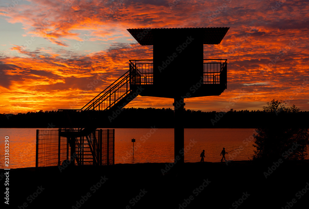 Two Boys Fishing Walk Past Lifeguard Tower At Lake Under Red Sky
