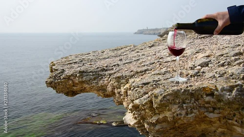 Red wine is poured into a glass on a background of rocky sea shore. A hand with a bottle pours wine into a glass standing on a rocky seashore 