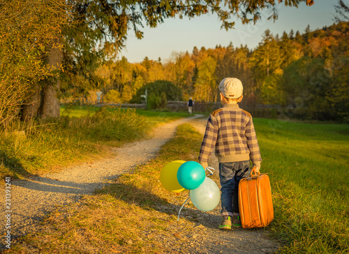 Happy little boy playing on road at sunset. He standing in the field and holding in hands suitcase and balloons.