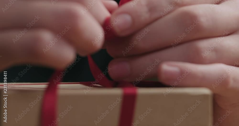 Slow motion macro shot of young female hands making red ribbon bow on craft paper gift box Slow motion macro shot of young female hands making red ribbon bow on craft paper gift box