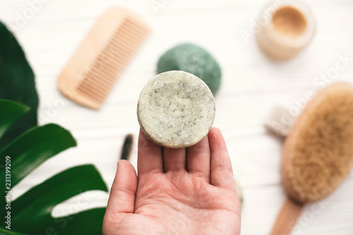 Hand holding natural solid shampoo bar on background of bamboo brush, deodorant, sponge on white wood with green monstera leaves. Zero waste. Choice plastic free eco products photo