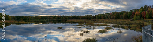 Puffy blue and white clouds are reflected in the still, mirror-like surface of a Virginia marshland. Meanwhile, green, orange, and red leaves adorn the trees in a surrounding forest. 