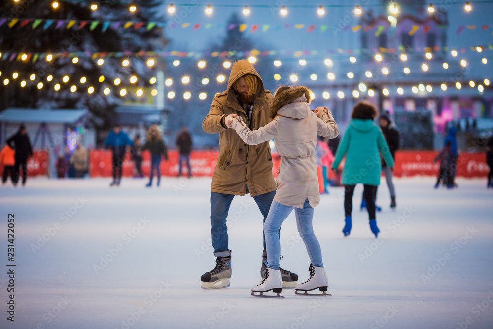 Young couple in love Caucasian man with blond hair with long hair and beard and beautiful woman have fun, active date skating on ice scene in town square in winter on Christmas Eve - obrazy, fototapety, plakaty 