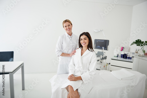 Portrait of charming woman in white bathrobe sitting on daybed while beautician touching her shoulder. They looking at camera and smiling photo