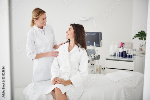 Portrait of charming woman in white bathrobe sitting on daybed and looking at beautician with smile while she touching her shoulder photo