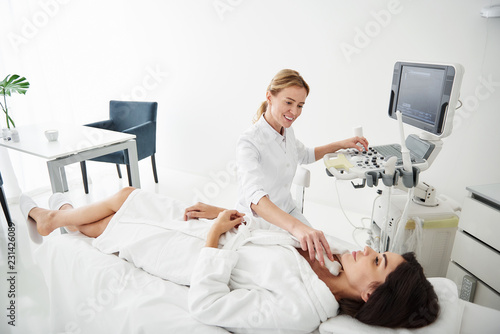 Portrait of attractive young woman in white bathrobe lying on daybed during medical examination while physician looking at her with smile