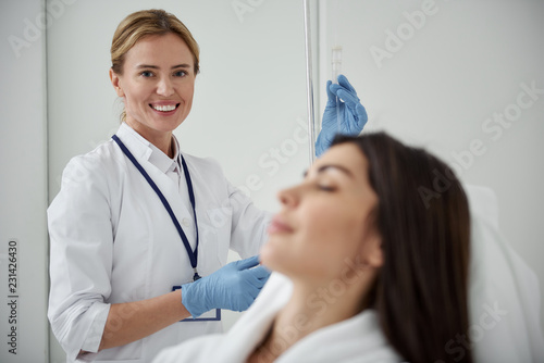 Waist up portrait of attractive woman in white lab coat regulating IV infusion. She is looking at camera and smiling