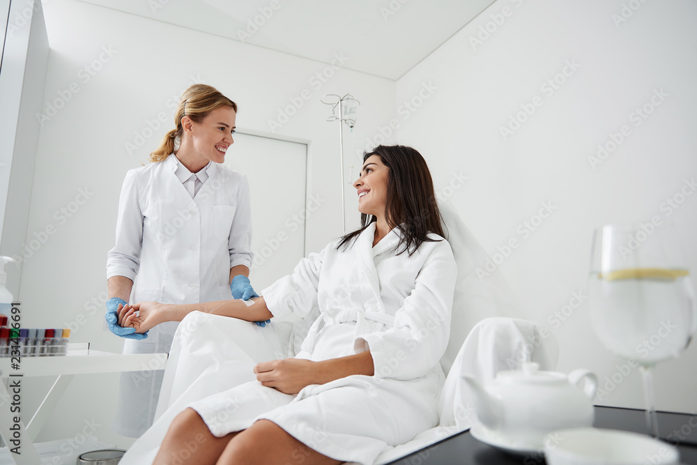 Fototapeta premium Side view portrait of charming woman in white bathrobe sitting in armchair and receiving IV infusion. She is looking at physician and smiling