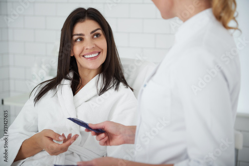 Portrait of charming lady in white bathrobe lying on daybed and looking at beautician with smile while she recommending effective skin care product