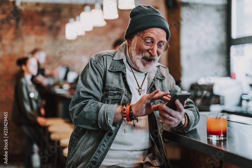 Waist up portrait of cheerful hipster pensioner sitting in bar and using smartphone