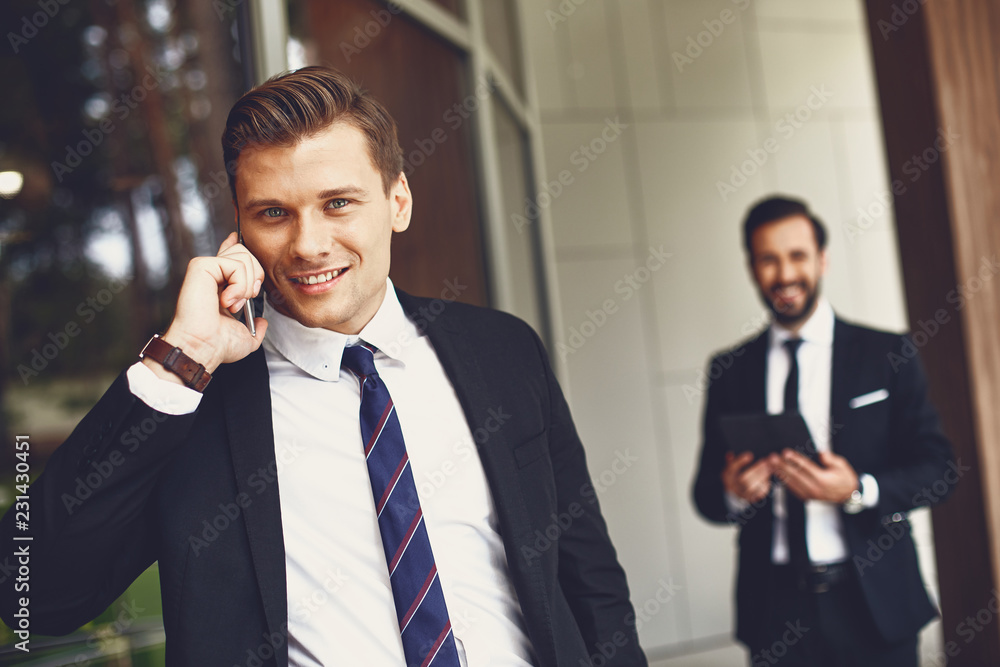 Attractive young man in elegant dark suit talking on the phone while his colleague standing not far from him Attractive young man in elegant dark suit talking on the phone while his colleague standing not far from him