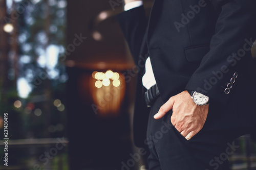 Atrakcyjny młody człowiek w ciemnym garniturze i zegarku na nadgarstku, który wkłada rękę do kieszeni spodni
