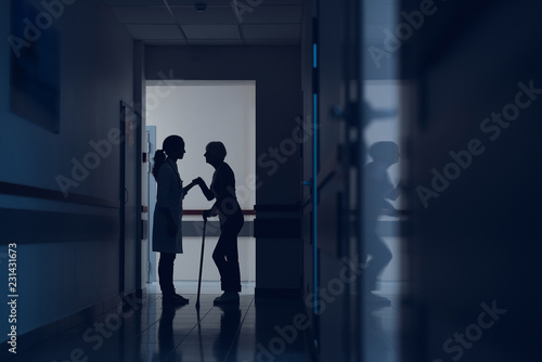 Woman and physician are standing in hospital corridor. Patient is walking with stick while female practitioner is holding her hand. Copy space in right side