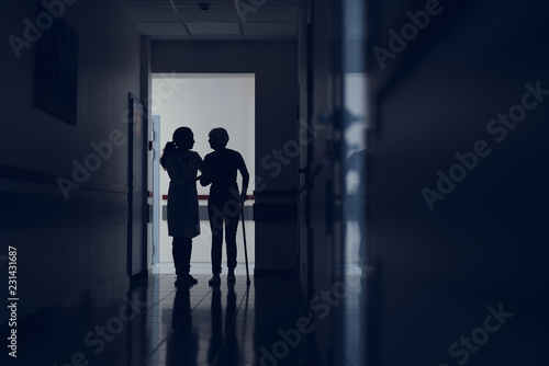 Woman is using stick for moving in clinic. Attentive practitioner is supporting her by hand while walking in corridor. Copy space in right side