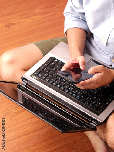 Closeup of a man sitting typing on his mobilephone with his laptop on his lap.