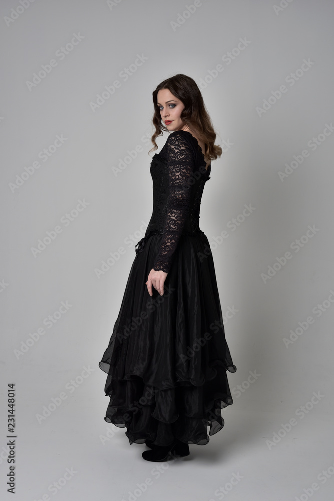 full length portrait of brunette girl wearing long black lace gown with corset. standing pose with back to the camera, grey studio background. full length portrait of brunette girl wearing long black lace gown with corset. standing pose with back to the camera, grey studio background.