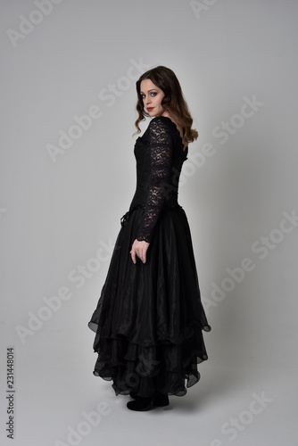 full length portrait of brunette girl wearing long black lace gown with corset. standing pose with back to the camera, grey studio background.