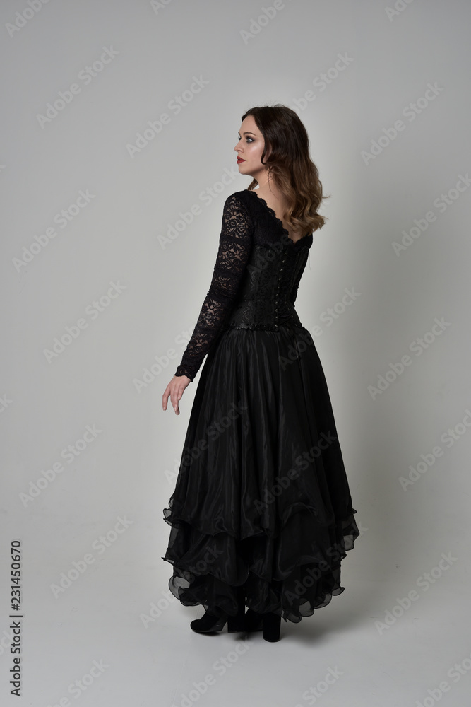 full length portrait of brunette girl wearing long black lace gown with corset. standing pose with back to the camera, grey studio background. full length portrait of brunette girl wearing long black lace gown with corset. standing pose with back to the camera, grey studio background.