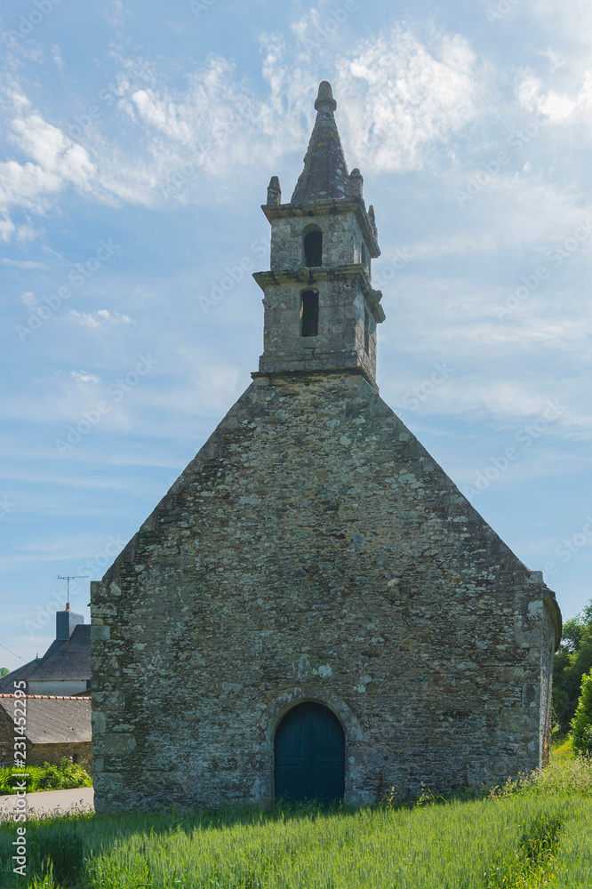 Small stone church in rural Brittany, France, 2017. Green grass in front and tiny blue entrance door, with clouds in blue sky behind