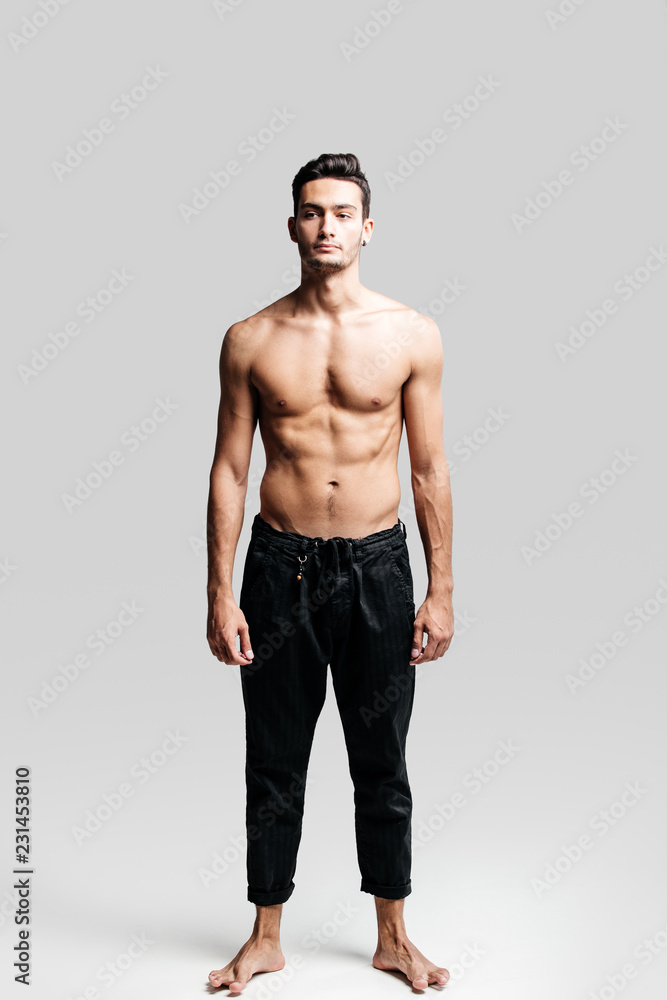 Fototapeta premium Dark-haired handsome young dancer with bare torso wearing a black sports pants is standing on a white background