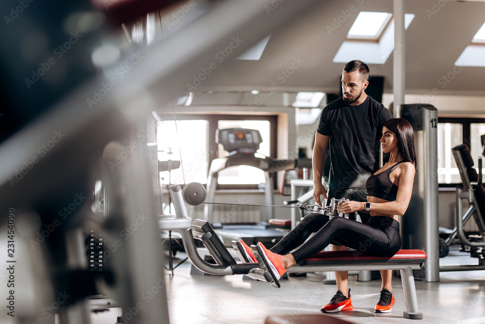 Beautiful slim girl dressed in black sport clothes is doing exercises for the abdominals on a special exercise machine under the supervision of a coach in the gym Beautiful slim girl dressed in black sport clothes is doing exercises for the abdominals on a special exercise machine under the supervision of a coach in the gym