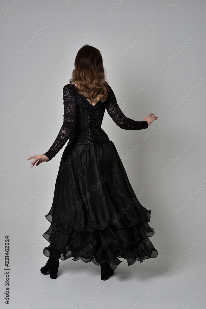 full length portrait of brunette girl wearing long black lace gown with corset. standing pose with back to the camera, grey studio background. full length portrait of brunette girl wearing long black lace gown with corset. standing pose with back to the camera, grey studio background.