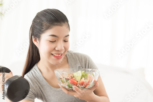 Young asian woman holding bowl of salad with dumbell at home. Fitness and healthy lifestyle concept