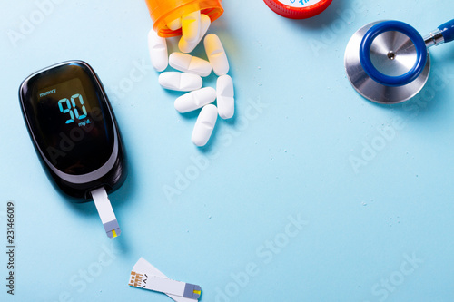 White pills in orange bottle with blood glucose meter on blue background with copy space