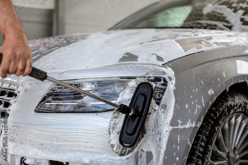 Front light of silver car washed in self serve carwash  man hand holding brush   cleaning the glass with shampoo