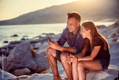 Father and daughter sitting on a rocky beach and using smart phone