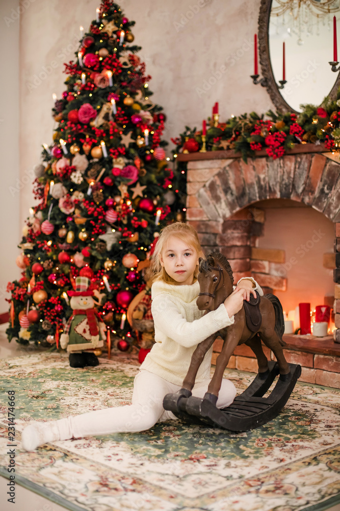 Christmas portrait of happy blonde child girl in white sweater siting on the floor near the Christmas tree and wooden toy horse. New Year Holidays