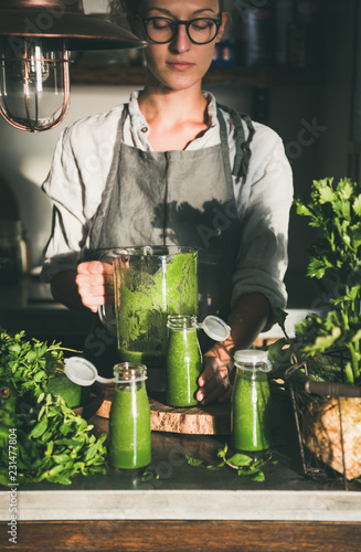 Making green detox take-away smoothie. Woman in linen apron and glasses pouring green smoothie drink from blender to bottle surrounded with vegetables and greens. Healthy, weight loss food concept