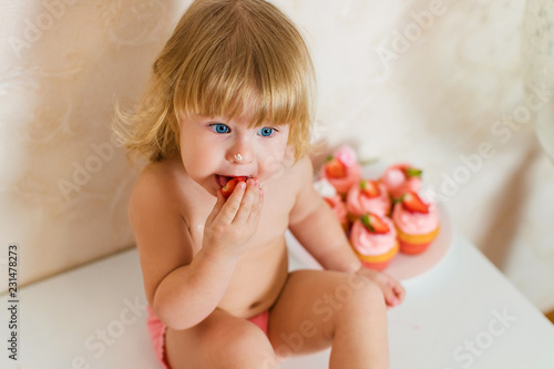 Little blonde baby girl two years old in pink pants sitting on the white table near her birthday cake and different pink sweets on the table