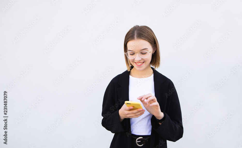Happy office worker woman standing on a white background and uses the smartphone, looking at screen smartphone and smiles. Portrait of a young teacher with a smartphone in his hands. Happy office worker woman standing on a white background and uses the smartphone, looking at screen smartphone and smiles. Portrait of a young teacher with a smartphone in his hands.