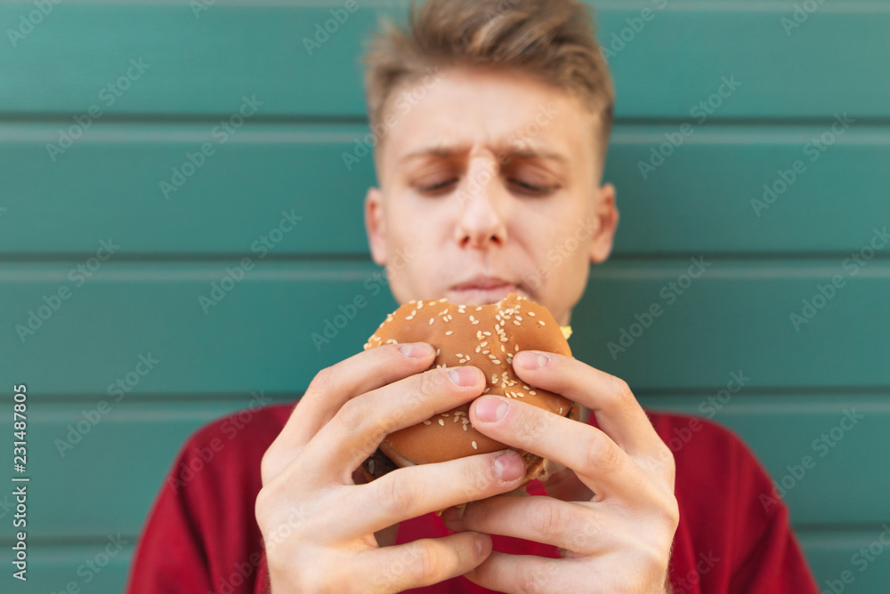 Close-up portrait of a beautiful young man with an appetizing burger in his hands on a turquoise background. Focus on the burger. Young man eats fast food on the street. Close-up portrait of a beautiful young man with an appetizing burger in his hands on a turquoise background. Focus on the burger. Young man eats fast food on the street.