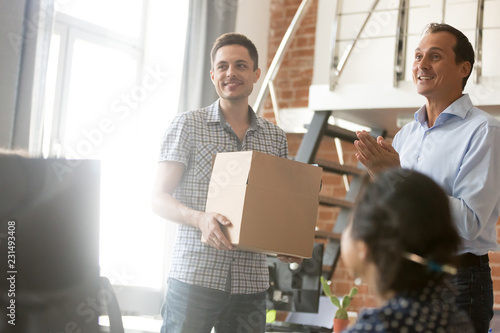 Excited leader boss introducing new employee holding box on first day at work, friendly ceo and team welcoming just hired member newcomer getting acquainted with male coworker, introduction concept