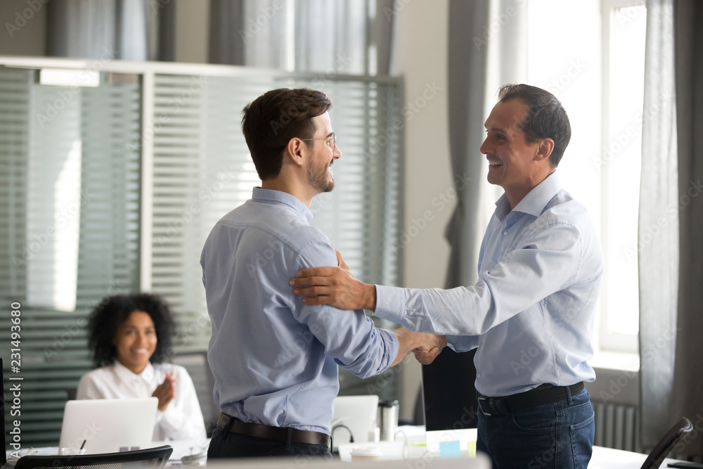 Smiling middle-aged ceo promoting motivating worker shaking hands congratulating with achievement promising respect bonus thanking for good work, team applauding, employee reward recognition concept Smiling middle-aged ceo promoting motivating worker shaking hands congratulating with achievement promising respect bonus thanking for good work, team applauding, employee reward recognition concept