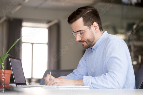 Focused business man making notes planning marketing economic research work with laptop, serious male student or worker writing studying online learning course preparing for test exam with computer