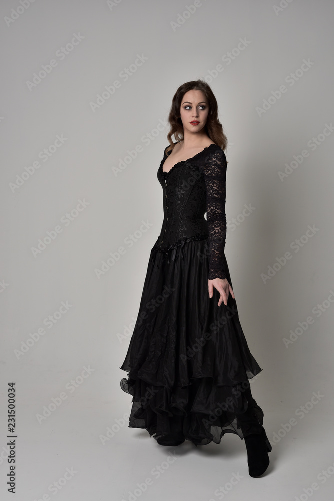 full length portrait of brunette girl wearing long black gown with corset. standing pose on grey studio background.