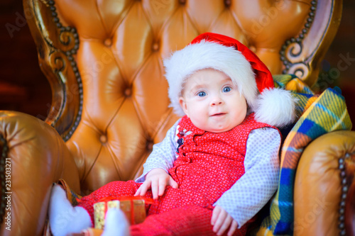 funny baby in a santa claus hat sitting in a chair with a small gift and smiling