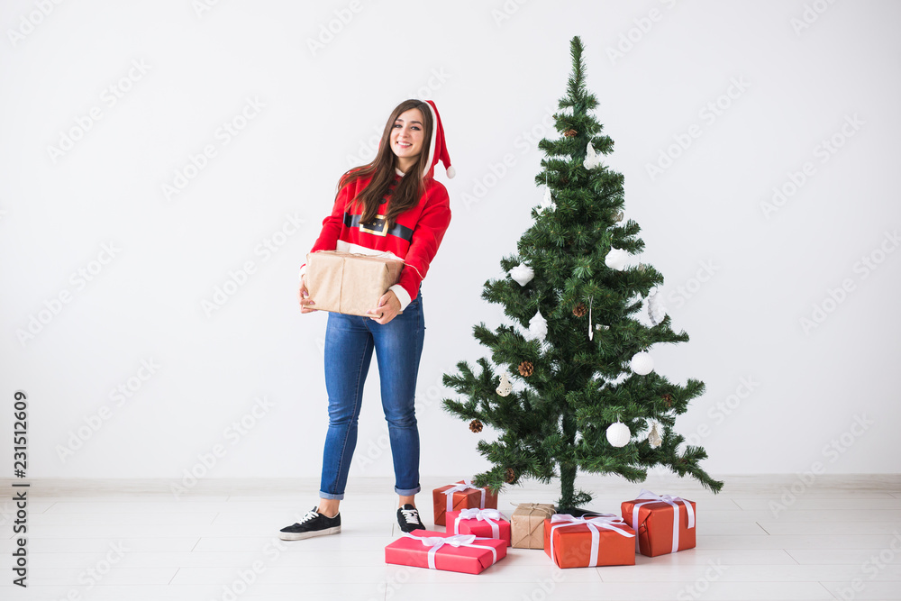 Young cheerful woman holding gift box wearing Santa costume. Christmas tree background with copy space Young cheerful woman holding gift box wearing Santa costume. Christmas tree background with copy space
