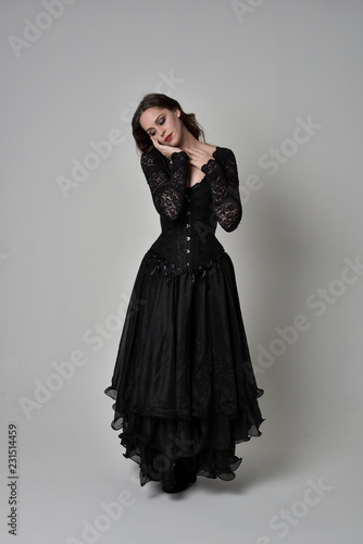 full length portrait of brunette girl wearing long black gown with corset. standing pose on grey studio background.