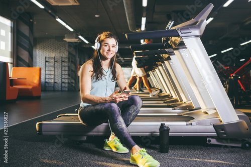 Beautiful young Caucasian girl sportswoman sitting, resting after training on treadmill against the backdrop of gym in sunny weather. Listens to music in sick white headphones,in hand holds the phone photo