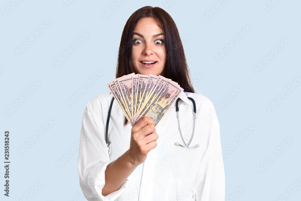 Shocked female doctor holds dollar bills, being rich, involved in corruption, cant believe she has much money, makes business on patients, poses against light blue background. Medicine is paid Shocked female doctor holds dollar bills, being rich, involved in corruption, cant believe she has much money, makes business on patients, poses against light blue background. Medicine is paid